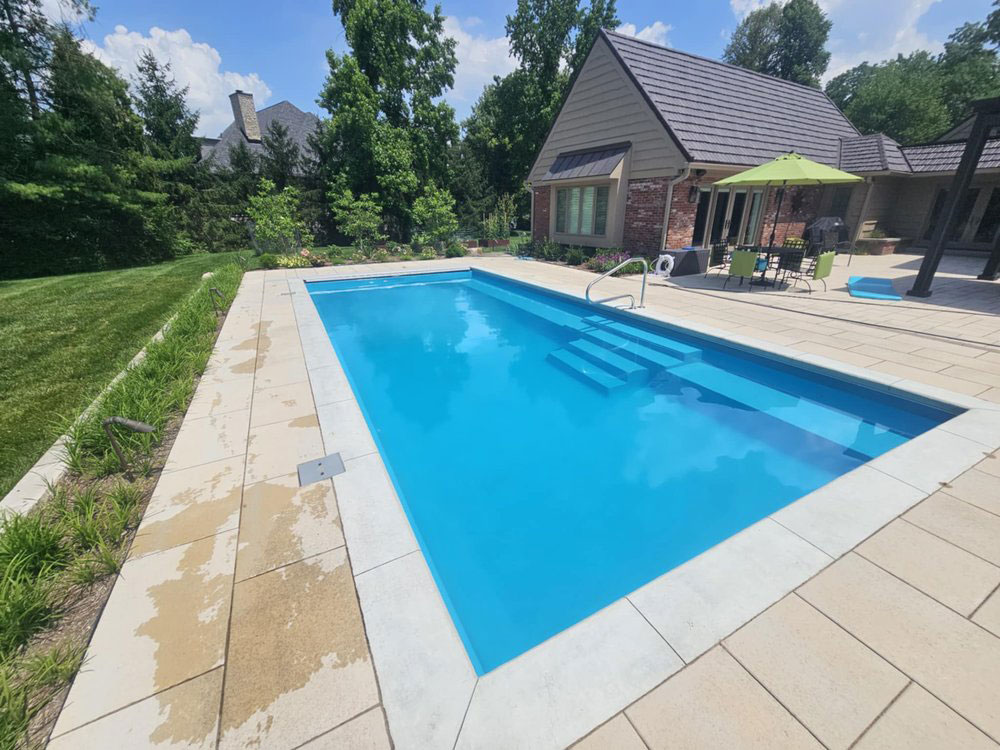 Rectangular fiberglass pool with sleek built-in steps and light stone patio in a landscaped backyard