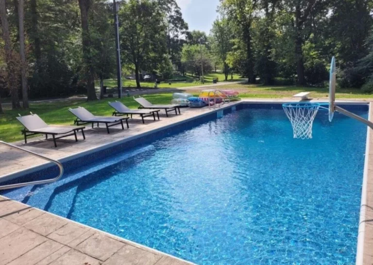 Rectangular vinyl liner pool with basketball hoop and lounge chairs in a wooded backyard